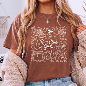 Op de afbeelding: Bruin t-shirt met een wit grafisch ontwerp met hardloopschoenen, een waterfles, shorts, een vest, een medaille en de tekst "Run Club Girlie". Het ontwerp bevat ook een startnummer en een hardloophorloge.