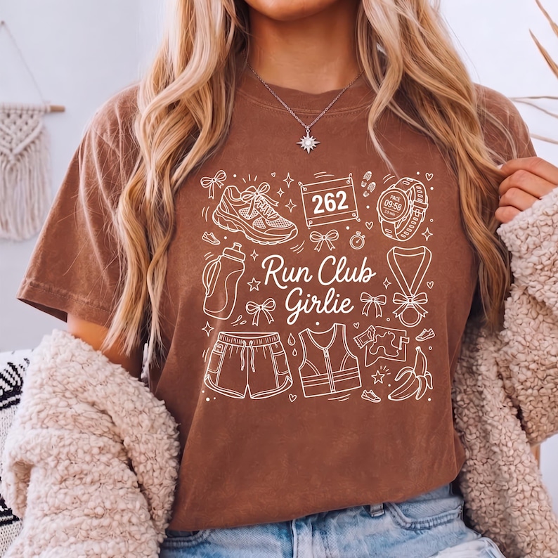 Op de afbeelding: Bruin t-shirt met een wit grafisch ontwerp met hardloopschoenen, een waterfles, shorts, een vest, een medaille en de tekst "Run Club Girlie". Het ontwerp bevat ook een startnummer en een hardloophorloge.