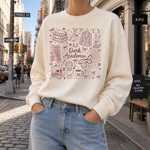 以下が含まれることがあります： クリーム色のスウェットシャツ。マルーン色のグラフィックには、本、ペン、ジャケット、ろうそく、スカル、そして「Dark Academia」の文字が描かれています。デザインにはコーヒーカップと手紙も含まれています。モデルは青いジーンズを着用しています。