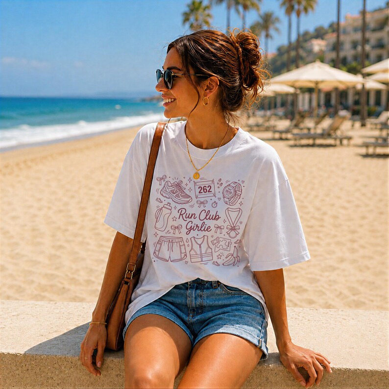 Op de afbeelding: Wit T-shirt met een paars grafisch ontwerp met hardloopiconen en de tekst "Run Club Girlie". Het shirt wordt gecombineerd met een blauwe spijkerbroek. De persoon draagt een zonnebril en een gouden ketting.