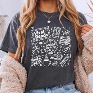 May include: Dark gray t-shirt with white graphic elements related to reading and social media. The design includes the text "Viral Reads," "Booktok made me do it," and "Must Read," along with icons of books, a phone, and a coffee cup.