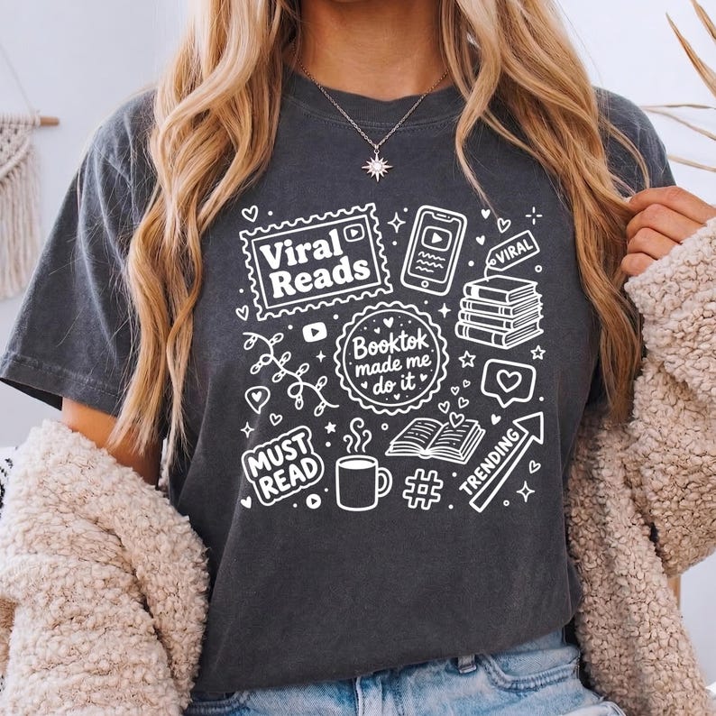 May include: Dark gray t-shirt with white graphic elements related to reading and social media. The design includes the text "Viral Reads," "Booktok made me do it," and "Must Read," along with icons of books, a phone, and a coffee cup.