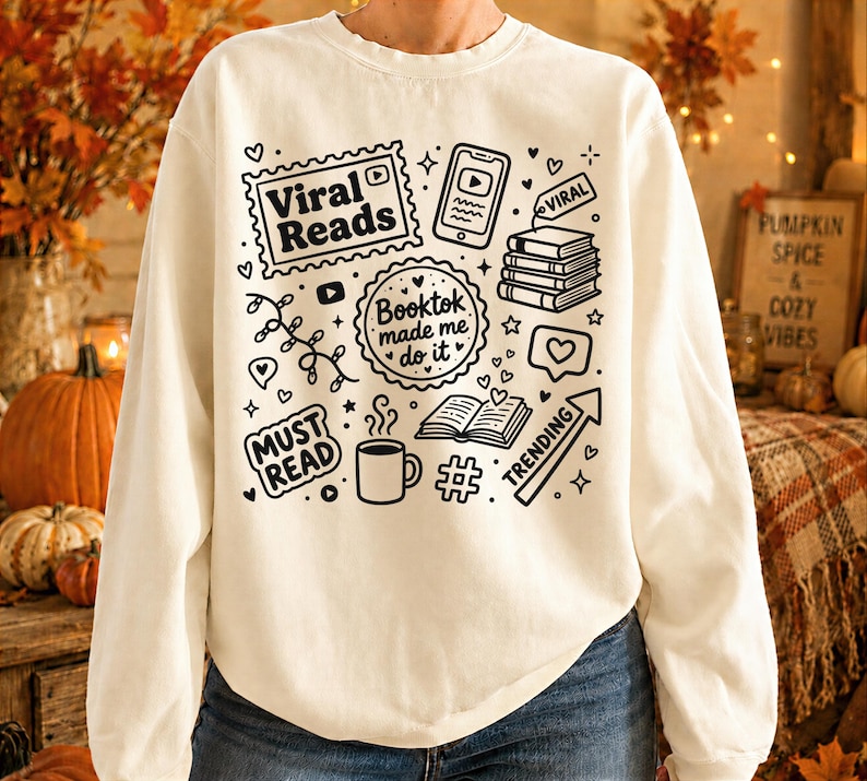 May include: Cream-colored sweatshirt with black graphic illustrations related to reading and social media. The design includes the text "Viral Reads," "Booktok made me do it," and "Must Read," along with icons of books, a phone, and a coffee cup.