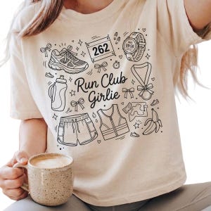 Op de afbeelding: Beige t-shirt met een zwart grafisch ontwerp met hardloopgerelateerde illustraties, waaronder een schoen, waterfles, shorts en een horloge. De tekst "Run Club Girlie" is ook in het ontwerp opgenomen.