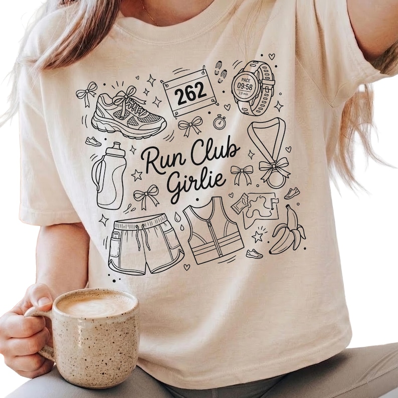 Op de afbeelding: Beige t-shirt met een zwart grafisch ontwerp met hardloopgerelateerde illustraties, waaronder een schoen, waterfles, shorts en een horloge. De tekst "Run Club Girlie" is ook in het ontwerp opgenomen.