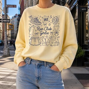 Op de afbeelding: Een lichtgeel sweatshirt met een marineblauw grafisch ontwerp. Het ontwerp bevat hardloopgerelateerde iconen en de tekst "Run Club Girlie". Het ontwerp toont een hardloopschoen, een korte broek, een waterfles, een horloge en een medaille.