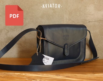 Puede incluir: Un bolso bandolera de cuero azul oscuro con cierre de solapa y un pequeño bolsillo en la parte delantera. El bolso tiene una correa de cuero marrón y una etiqueta que dice "AVIATOR".