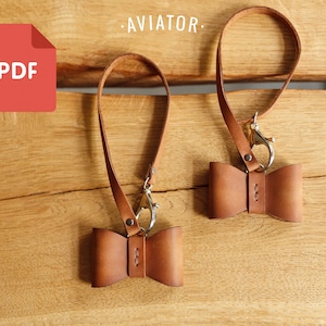 May include: Two brown leather bow tie keychains with silver clasps and straps. The keychains are hanging on a wooden surface. The text "AVIATOR" is visible above the keychains.