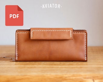 Puede incluir: Una cartera de cuero marrón con cierre de solapa y costuras blancas. La cartera está etiquetada como "AVIATOR."