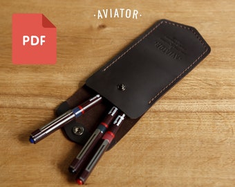 Puede incluir: Un portaplumas de cuero marrón con la palabra "AVIATOR" grabada en la parte delantera. Hay tres plumas dentro del portaplumas, dos rojas y una azul.