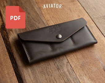 Puede incluir: Una cartera de cuero marrón con cierre de solapa y tres ranuras para tarjetas. La cartera tiene un cierre de botón plateado y el texto "AVIATOR" grabado en la parte delantera.