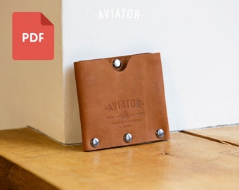 Puede incluir: Una cartera de cuero marrón con el texto "AVIATOR" grabado en la parte delantera. La cartera tiene remaches de metal plateado alrededor de los bordes.