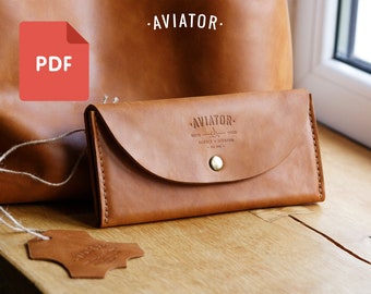 Puede incluir: Una cartera de cuero marrón con cierre de solapa y un botón de latón. La cartera tiene el texto "AVIATOR" grabado en la parte delantera. La cartera está sobre una superficie de madera junto a un bolso de cuero marrón y una etiqueta de cuero.