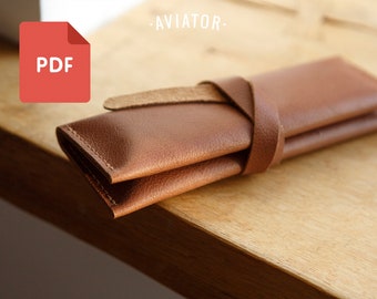 Puede incluir: Una funda de cuero marrón con cierre de correa. La funda está doblada por la mitad y la correa está envuelta alrededor de ella. La funda está sobre una superficie de madera. El texto "PDF" es visible en la esquina superior izquierda de la imagen.