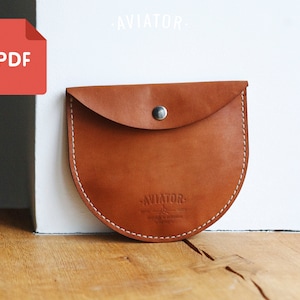 May include: A brown leather pouch with a curved bottom and a flap secured by a silver snap. White stitching outlines the edges. The word "AVIATOR" is stamped on the front. A red PDF icon is in the upper left corner.