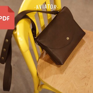 Könnte beinhalten: Eine braune Leder-Umhängetasche mit einer Klappe und einem goldenen Knopf. Die Tasche liegt auf einem gelben Stuhl mit dem Text "AVIATOR" auf der Rückenlehne.
