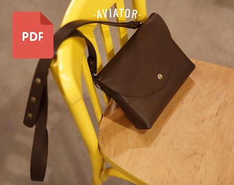 Puede incluir: Un bolso de cuero marrón cruzado con cierre de solapa y un botón dorado. El bolso está sentado en una silla amarilla con el texto "AVIATOR" impreso en el respaldo de la silla.