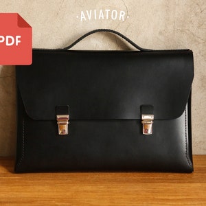 May include: Black leather briefcase with silver-tone clasps and a top handle. The word "AVIATOR" is above the briefcase. A red PDF icon is in the upper left corner. The briefcase is on a wooden surface.