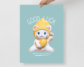 May include: A poster featuring a white cat figurine wearing a yellow lemon hat and holding a lemon. The words "GOOD LUCK" are written above the cat. The background is a light teal color.