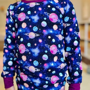 Könnte beinhalten: Ein langärmliges Shirt mit dunkelblauem Hintergrund und einem farbenfrohen Galaxie-Druck mit Planeten, Sternen und Nebeln. Das Shirt hat einen violetten Saum an Ausschnitt und Ärmelbündchen.