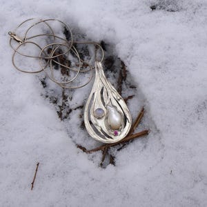 May include: A silver teardrop-shaped pendant necklace with a white pearl and a moonstone set in a delicate silver design. The necklace is lying on a bed of snow.