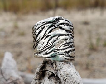 May include: A wide, silver ring with a textured, organic design. The ring's surface features a pattern of flowing lines and cut-out sections, creating a unique, artistic look. The ring is displayed on a piece of driftwood against a blurred natural background.
