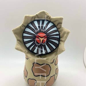 Pode incluir: Um brinquedo de pelúcia em forma de criatura com uma boca grande, redonda, listrada de marrom e branco com um centro vermelho. A criatura é bege com manchas marrons e tem um corpo arredondado.