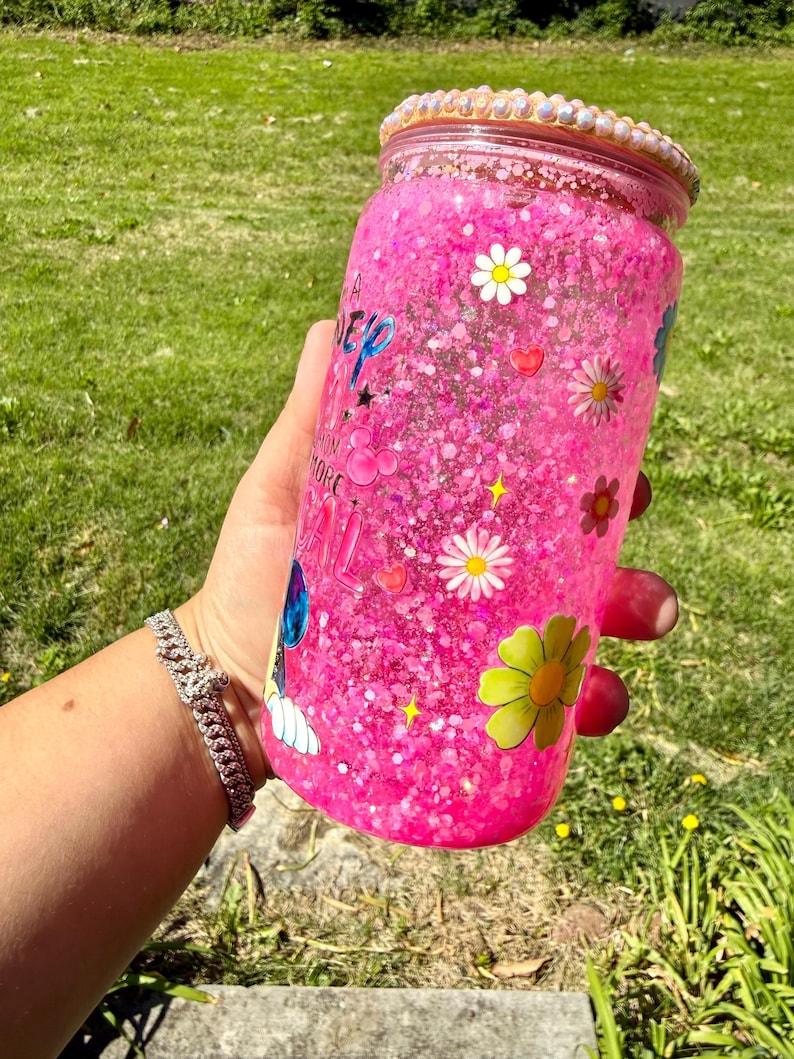 Op de afbeelding: Een roze glitterbeker met madeliefjes- en hartstickers. De beker heeft een met strass versierde rand en is gevuld met roze glitter. De beker heeft de tekst "A Step More" en een stripfiguur.