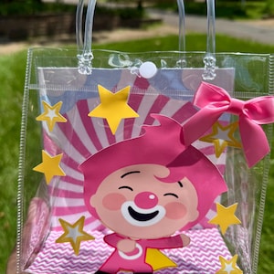 Puede incluir: Bolsa de mano de plástico transparente con un personaje de dibujos animados rosa, estrellas amarillas y un lazo rosa. La bolsa tiene asas transparentes y un cierre de botón blanco. El fondo presenta un diseño a rayas rosas y blancas.