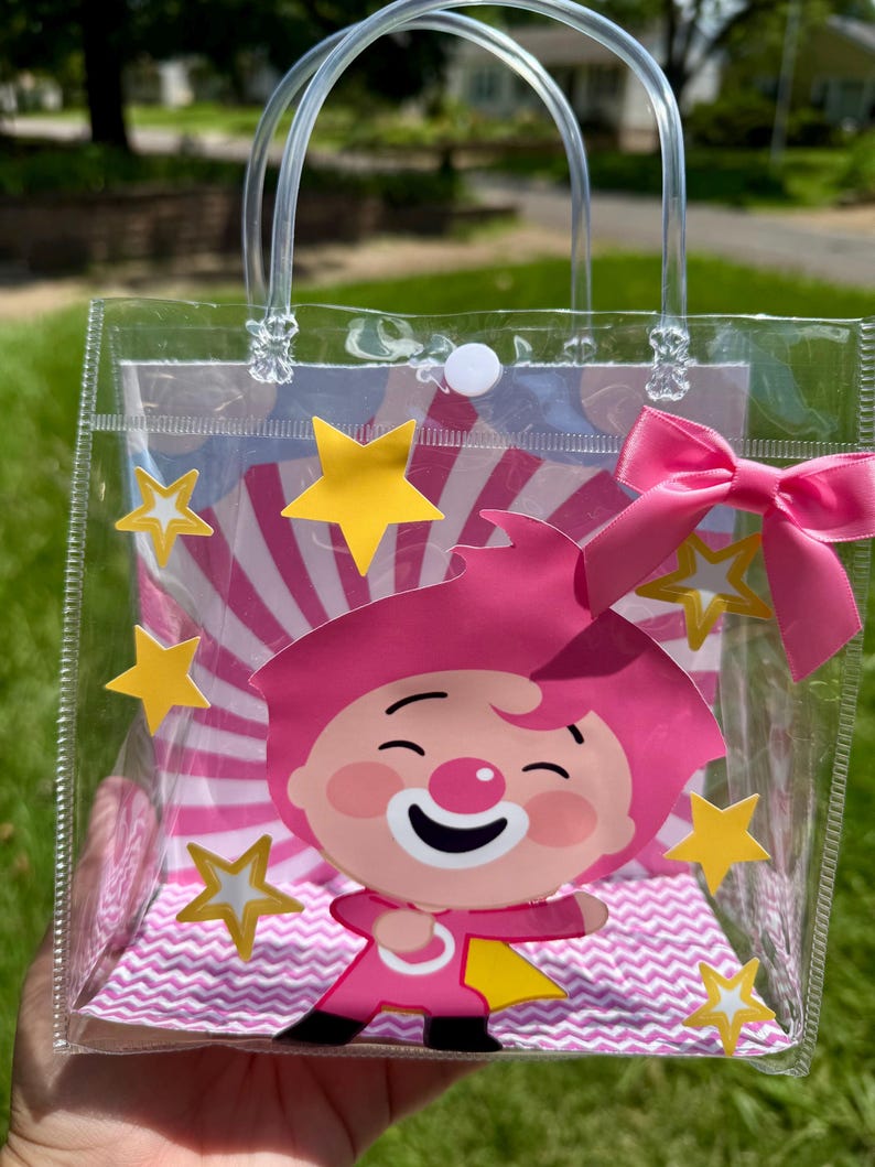 May include: Clear plastic tote bag with a pink cartoon character, yellow stars, and a pink bow. The bag has clear handles and a white button closure. The background features a pink and white striped design.
