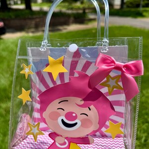 May include: A clear plastic tote bag featuring a pink clown character with a white nose and a pink bow. The bag has a clear handle and is decorated with yellow stars and a pink and white striped background.