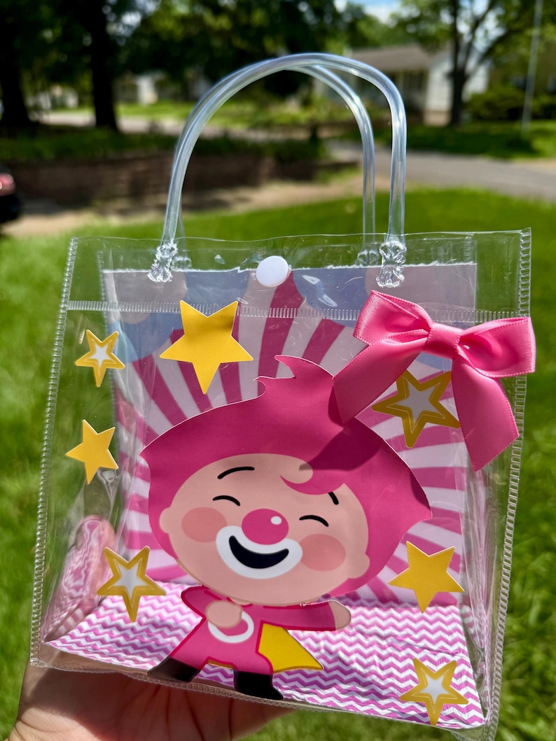 May include: A clear plastic tote bag featuring a pink clown character with a white nose and a pink bow. The bag has a clear handle and is decorated with yellow stars and a pink and white striped background.