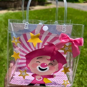 May include: Clear plastic tote bag with a cartoon clown graphic. The bag features a pink bow, yellow star accents, and a pink and white striped background. The bag has clear plastic handles.