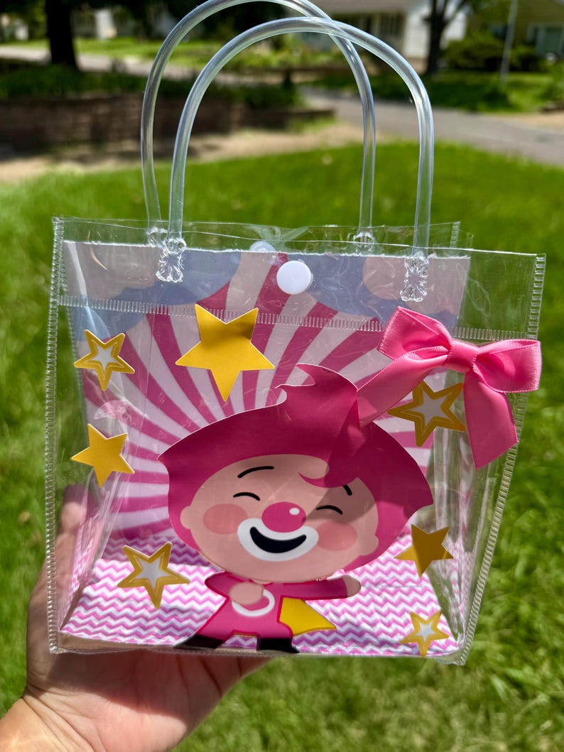 May include: Clear plastic tote bag with a cartoon clown graphic. The bag features a pink bow, yellow star accents, and a pink and white striped background. The bag has clear plastic handles.
