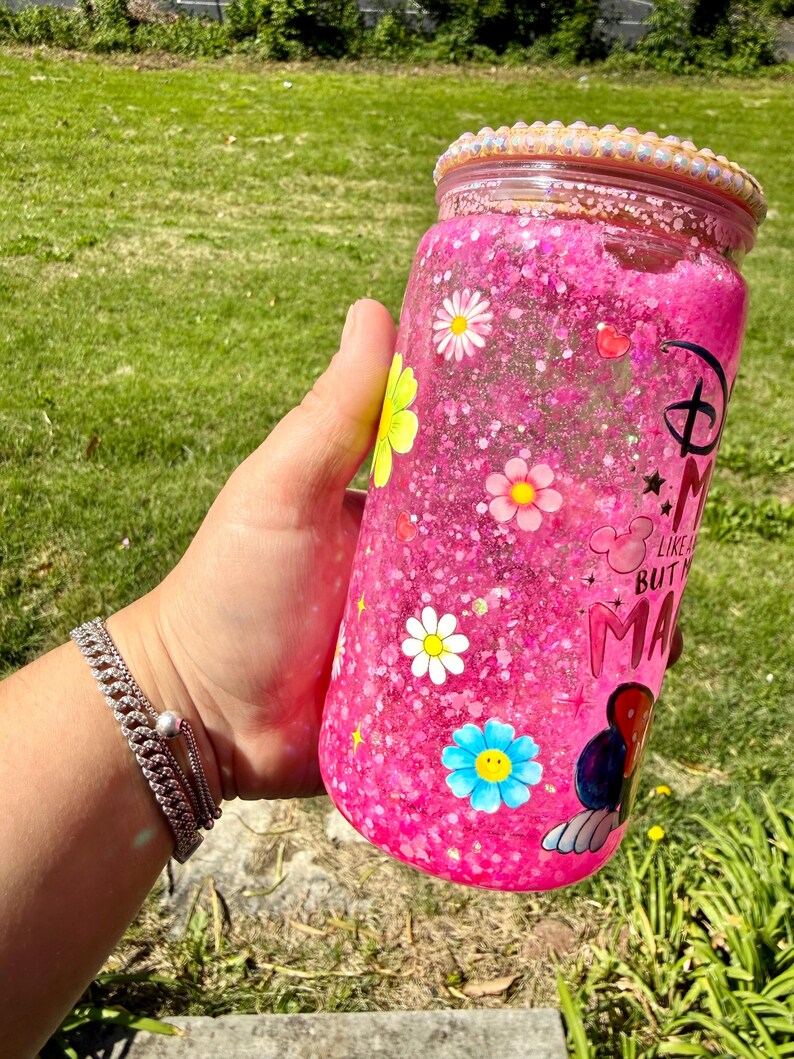 Op de afbeelding: Een roze glitterbeker met een bloemenontwerp en de tekst "Minnie Mouse Like a Boss But Make it Magic". De beker heeft een decoratieve rand en is gevuld met roze glitter en kleine bloemen.