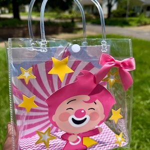 May include: Clear plastic tote bag featuring a cartoon character with pink hair and outfit, a white smiling face, and yellow stars. The bag has clear handles and a pink bow. The background has pink and white stripes.