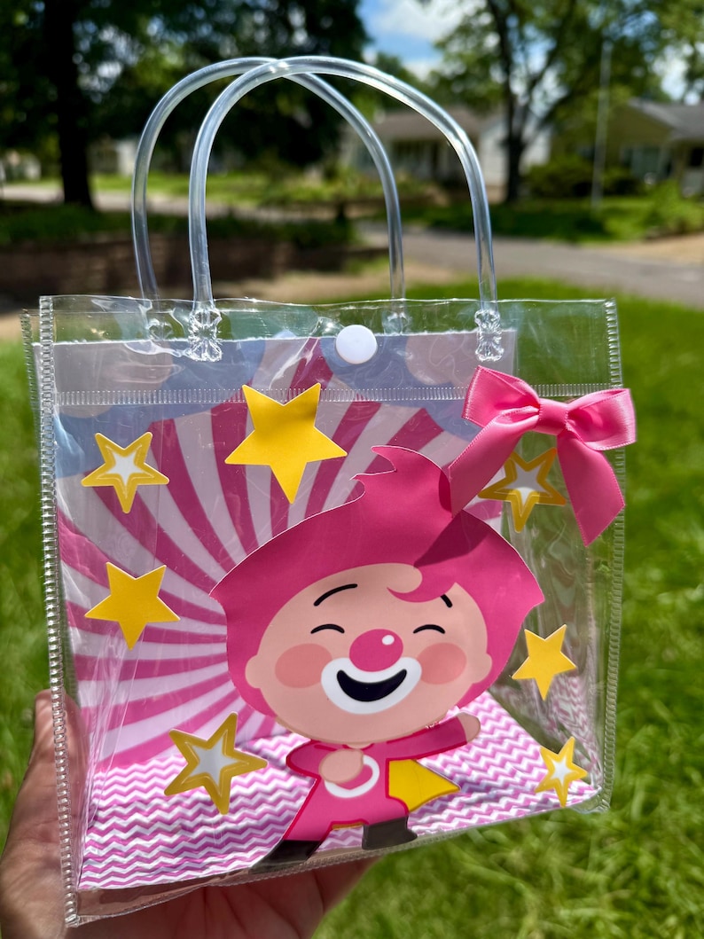 May include: Clear plastic tote bag featuring a cartoon character with pink hair and outfit, a white smiling face, and yellow stars. The bag has clear handles and a pink bow. The background has pink and white stripes.