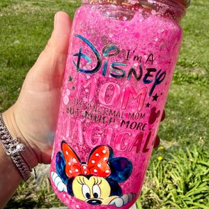 Op de afbeelding: Een roze, met glitter gevulde beker met de tekst "I'm a Disney Mom" en een Minnie Mouse-afbeelding. De beker heeft een decoratieve, met juwelen bezette rand en wordt door een persoon vastgehouden.