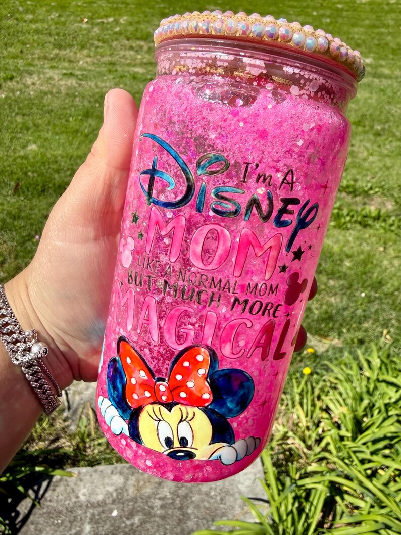 Op de afbeelding: Een roze, met glitter gevulde beker met de tekst "I'm a Disney Mom" en een Minnie Mouse-afbeelding. De beker heeft een decoratieve, met juwelen bezette rand en wordt door een persoon vastgehouden.