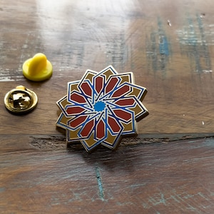 May include: A gold-toned pin with a red, blue, and gold geometric design. The pin is shaped like a flower with multiple points.