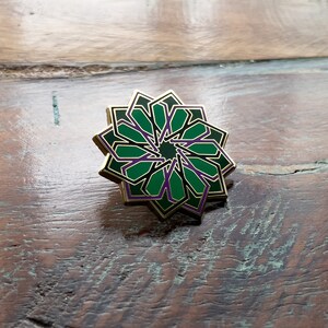 May include: A gold-colored pin with a green and purple geometric design. The design is a stylized starburst with multiple points.