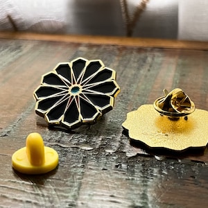 May include: A gold-toned enamel pin with a black and red geometric design. The pin features a circular shape with a starburst pattern. The back of the pin is gold-toned and has a pin back.