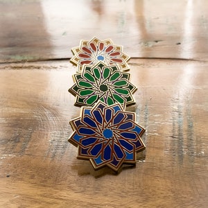 May include: Four gold-colored metal pins with intricate geometric designs. The pins are arranged in a row, with the top pin featuring a red and orange design, the second pin featuring a green and purple design, the third pin featuring a blue and purple design, and the bottom pin featuring a red and blue design.