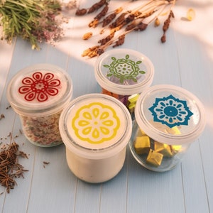 Op de afbeelding: Vier glazen potten met deksels. Elk deksel heeft een ander kleurrijk ontwerp. De potten zijn gevuld met verschillende inhoud.