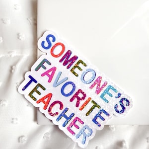 May include: A colorful sticker with the text "SOMEONE'S FAVORITE TEACHER" in various colors. The sticker has a white border and is placed on a white surface. The text is in a playful font.