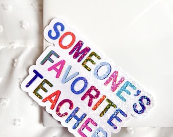 May include: A colorful sticker with the text "SOMEONE'S FAVORITE TEACHER" in various colors. The sticker has a white border and is placed on a white surface. The text is in a playful font.