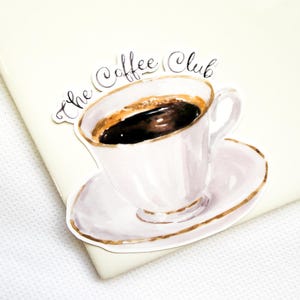 May include: A die-cut sticker featuring a watercolor illustration of a white teacup filled with black coffee on a matching saucer with gold trim. The text "The Coffee Club" is written in black cursive script above the cup.