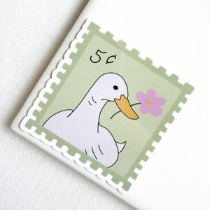 May include: A square sticker shaped like a postage stamp with a light green background. The design features a white duck with a yellow beak holding a small pink flower. The text 5c is printed in black in the upper left corner of the stamp.