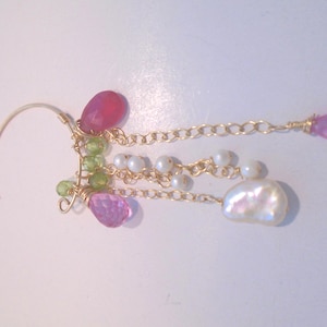 May include: A gold-toned ear cuff featuring a cluster of pink and green gemstones, small white pearls, and a large, irregular pearl. The design includes delicate gold chains and wirework, creating a unique and elegant accessory.