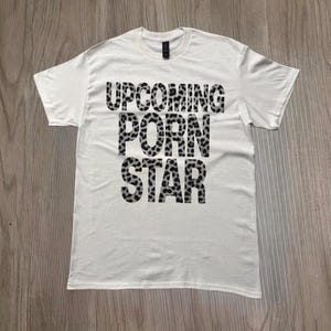 Puede incluir: Camiseta blanca con el texto "UPCOMING PORN STAR" en negro con estampado de leopardo. La camiseta tiene mangas cortas y cuello redondo. La prenda se muestra sobre una superficie de madera.
